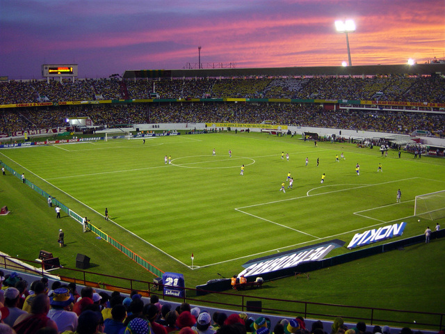 fotbalový stadion, stmívá se, růžová obloha, fotbalisti na hřišti, fanoušci všude po tribunách